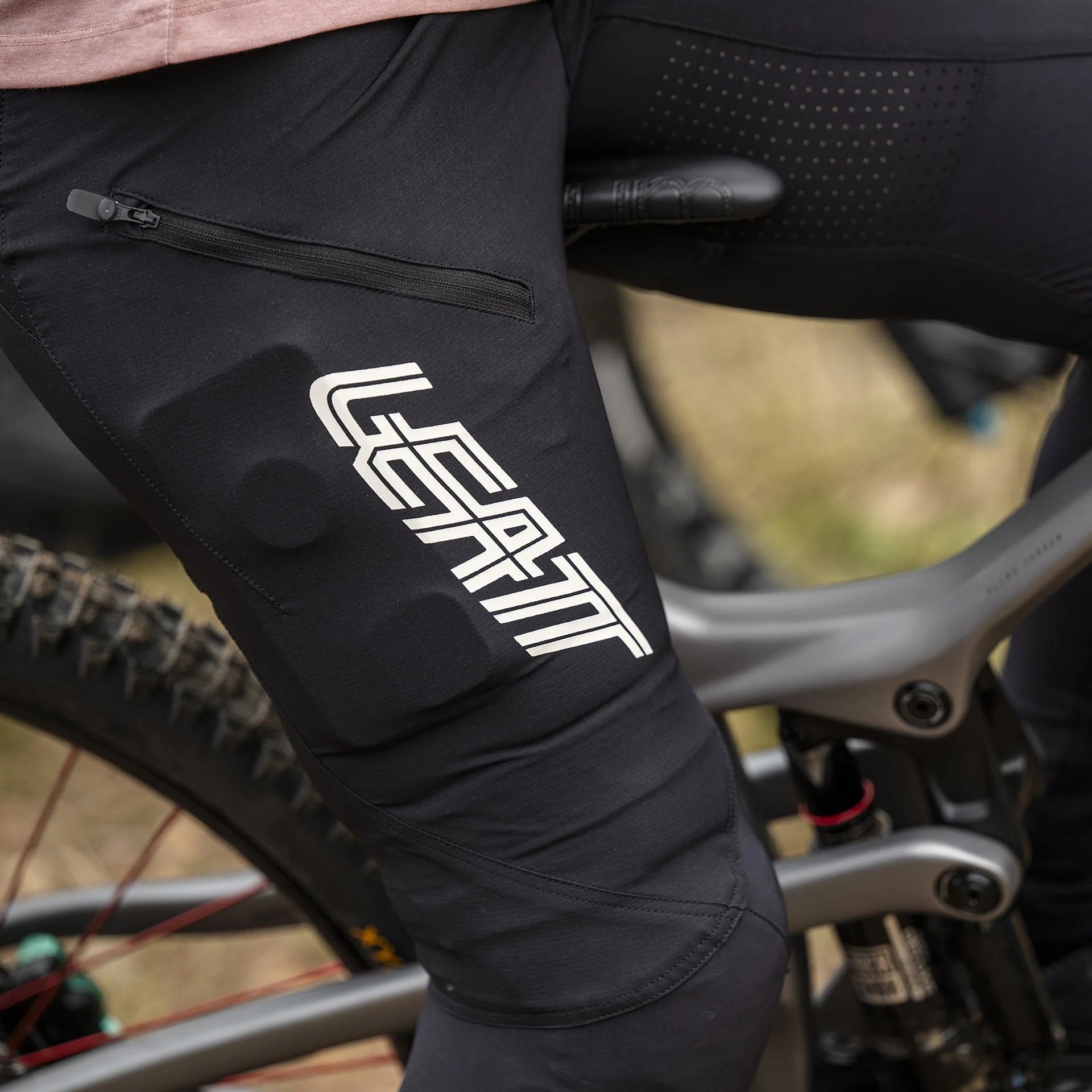 Person wearing Leatt MTB Gravity 3.0 black pants with a brand logo, sitting on a bicycle.