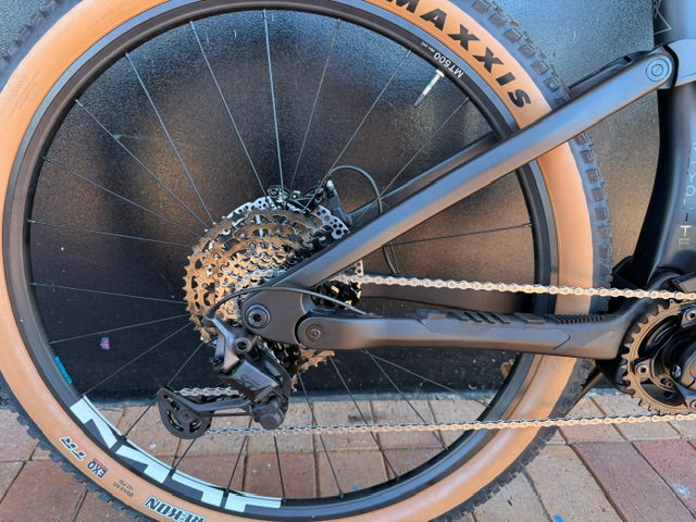Rhyolite Base+ Close-up of a bicycle rear wheel with chain and gear system on a paved surface.