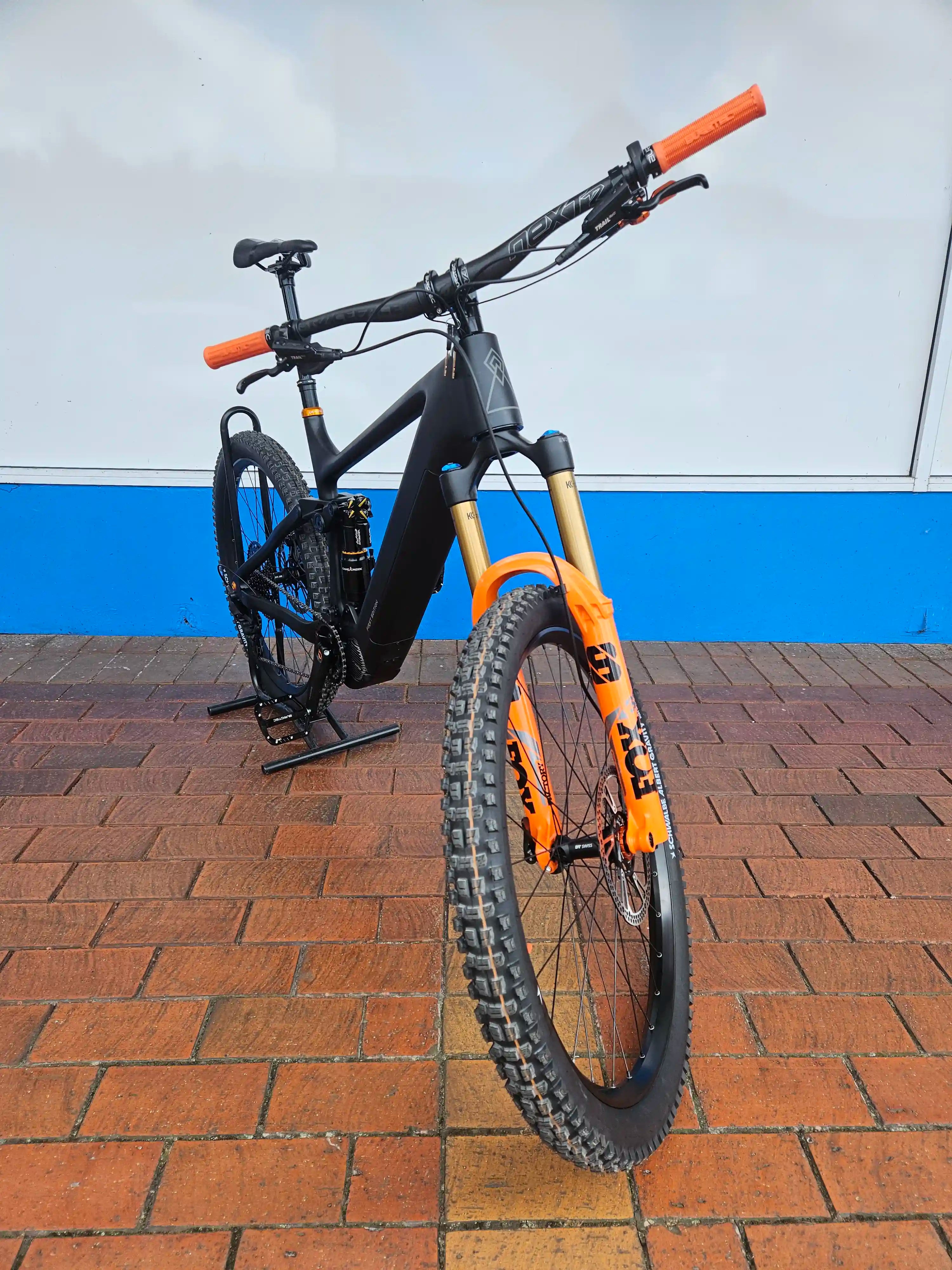 Orange and black Rhyolite Elite E-MTB leaning against a white and blue wall on a brick pavement.