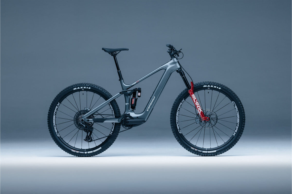 Tundra-10 gray full-suspension electric mountain bike with red front fork accents, black tires, and a visible Maxxis logo on the wheels, photographed in profile against a smooth gray studio background.