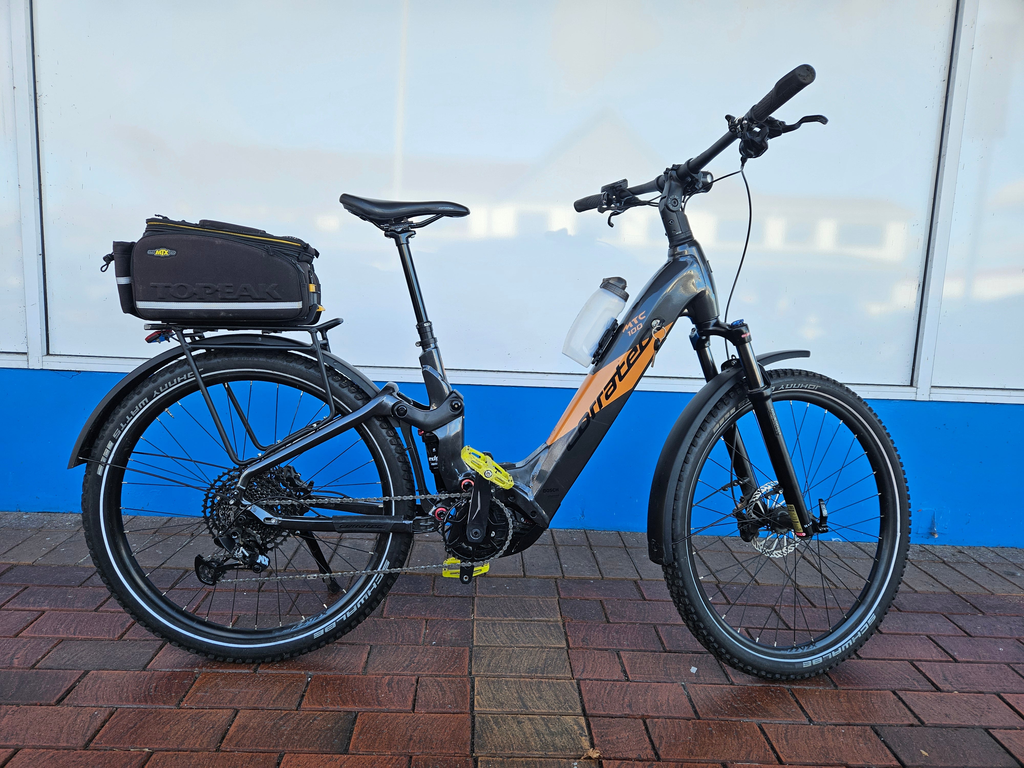 Electric bicycle with a black bag on a brick pavement against a blue and white wall.