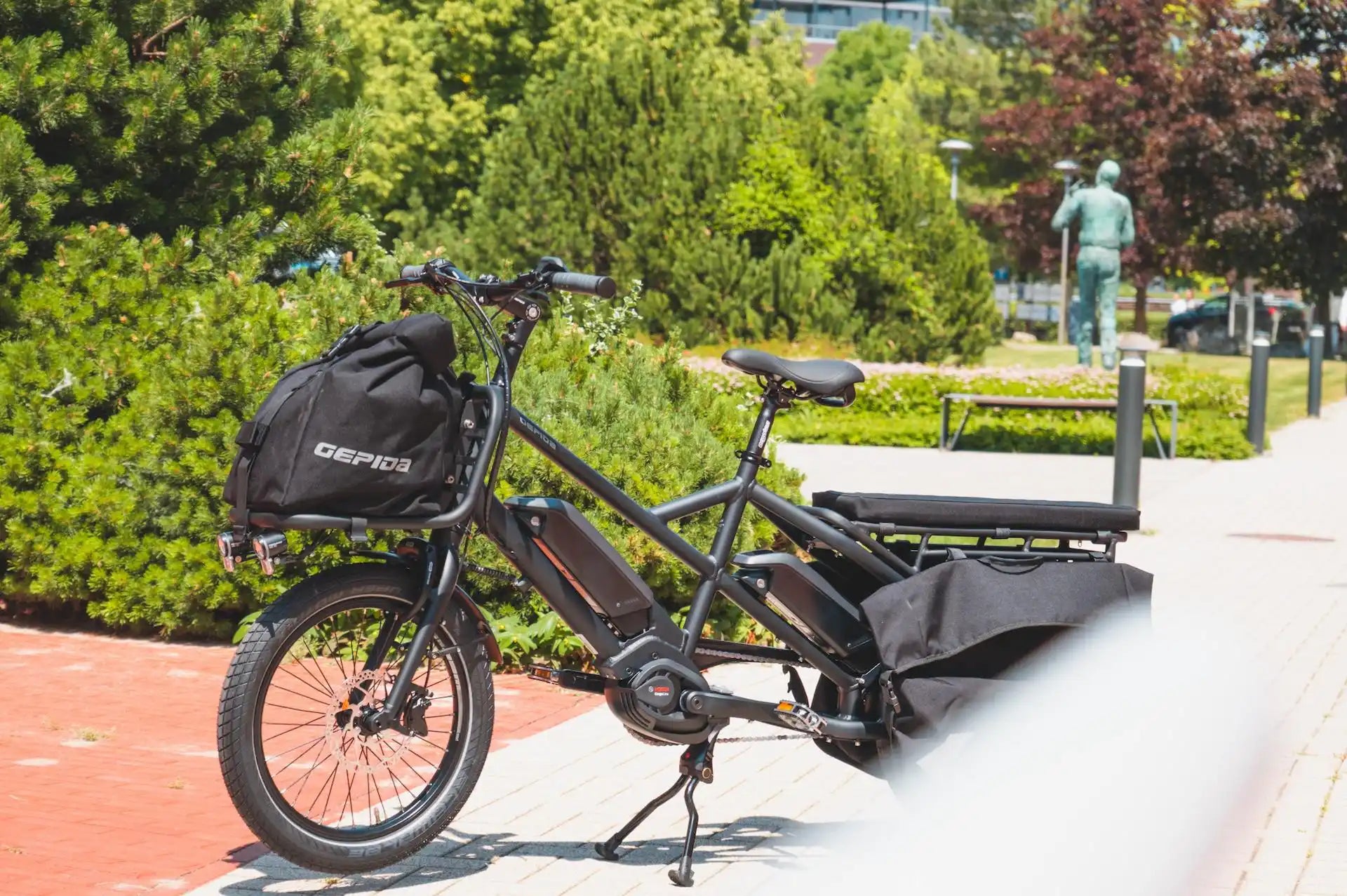 Gepida Cargo City E-Bike with a backpack on a sidewalk with greenery in the background