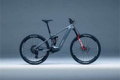 Tundra-10 gray full-suspension electric mountain bike with red front fork accents, black tires, and a visible Maxxis logo on the wheels, photographed in profile against a smooth gray studio background.