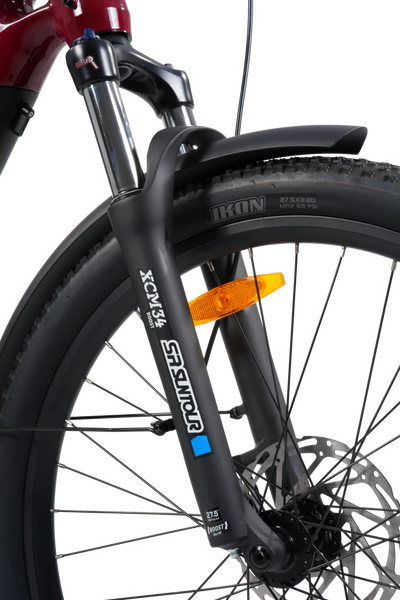 Close-up of a bicycle front wheel and suspension fork, showing a black knobby tire with an orange reflector and a black fork branded SCM with a blue accent.