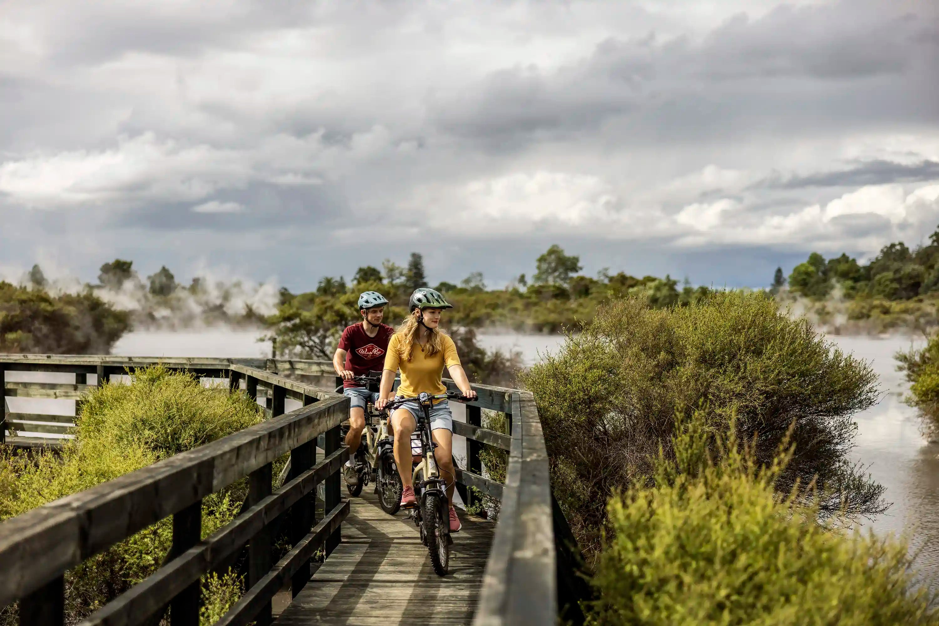 Two people cycling on rental E-bikes on a wooden bridge on Whaka E-bike Trail in Rotorua with mist and greenery.