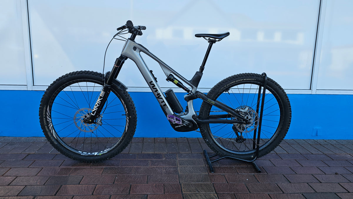 A full-suspension electric mountain bike with a matte gray frame and black components, displayed on a black stand against a blue and white wall.