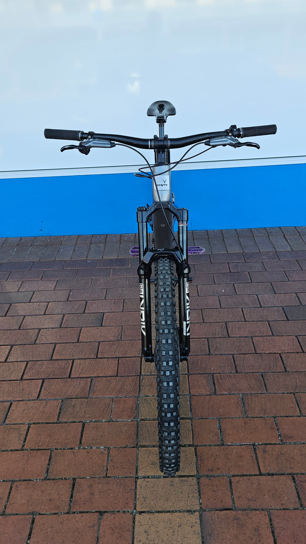 A black mountain bike with a front suspension fork and knobby tires, photographed from a head-on perspective on a brick surface with a blue and white wall in the background.
