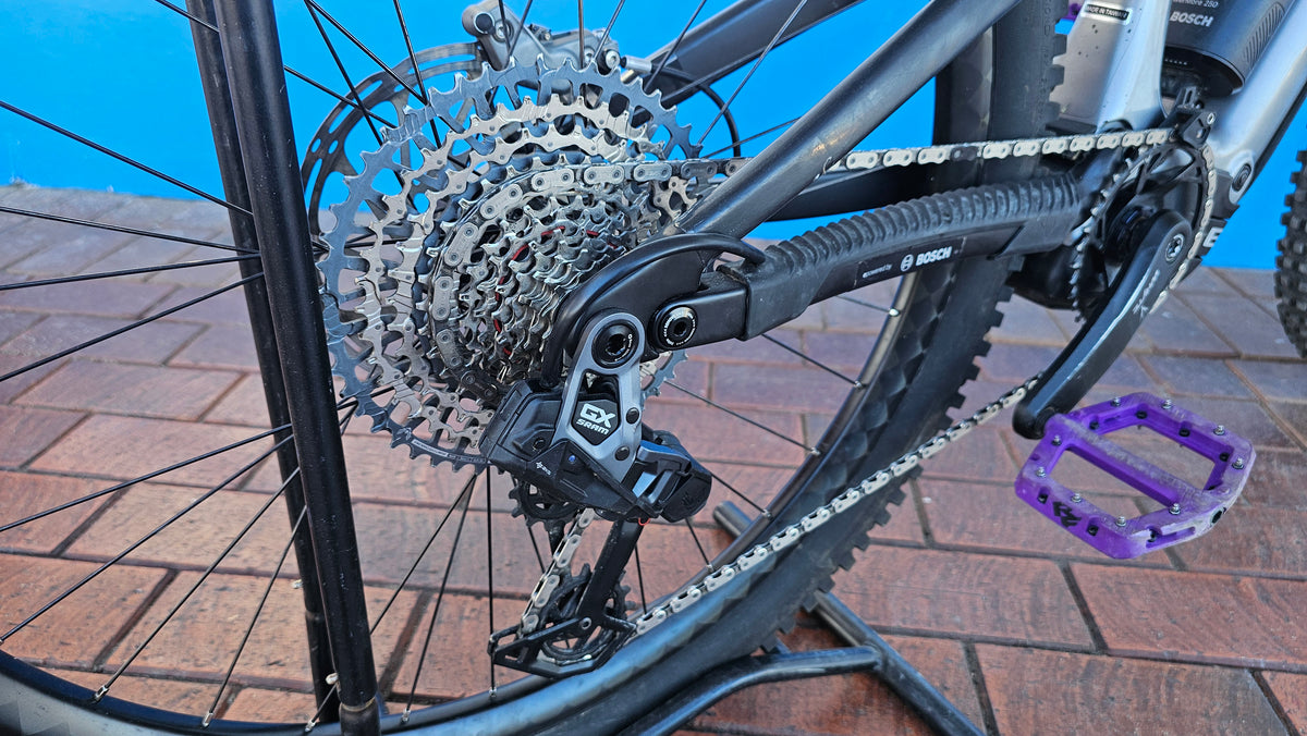Close-up of a mountain bike's rear wheel and drivetrain, showing a silver disc brake rotor, multi-gear cassette, chainring, and derailleur, with a purple pedal visible against a blue backdrop and brick floor.