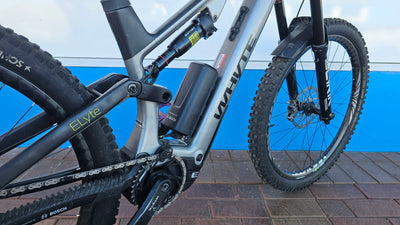 Close-up of a mountain bike frame and components, showing a silver and black frame with a visible 'WHITE' logo, rear suspension, rear wheel, and chainring area against a blue and white wall background.