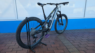 A silver and black full-suspension mountain bike with a battery mounted on the frame, displayed on a black stand on a brick surface in front of a blue and white wall.