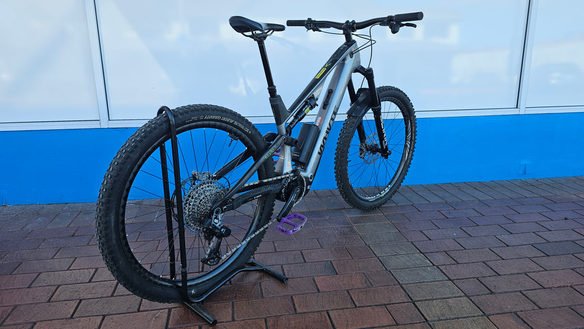 A silver and black full-suspension mountain bike with a battery mounted on the frame, displayed on a black stand on a brick surface in front of a blue and white wall.